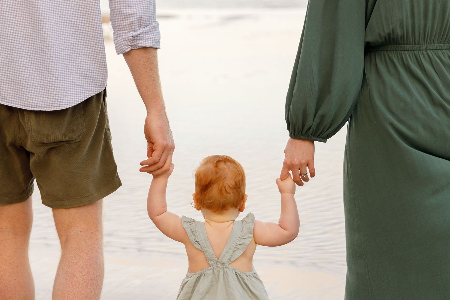 A toddler in a gray dress holds hands with adults while walking together on a bright path.