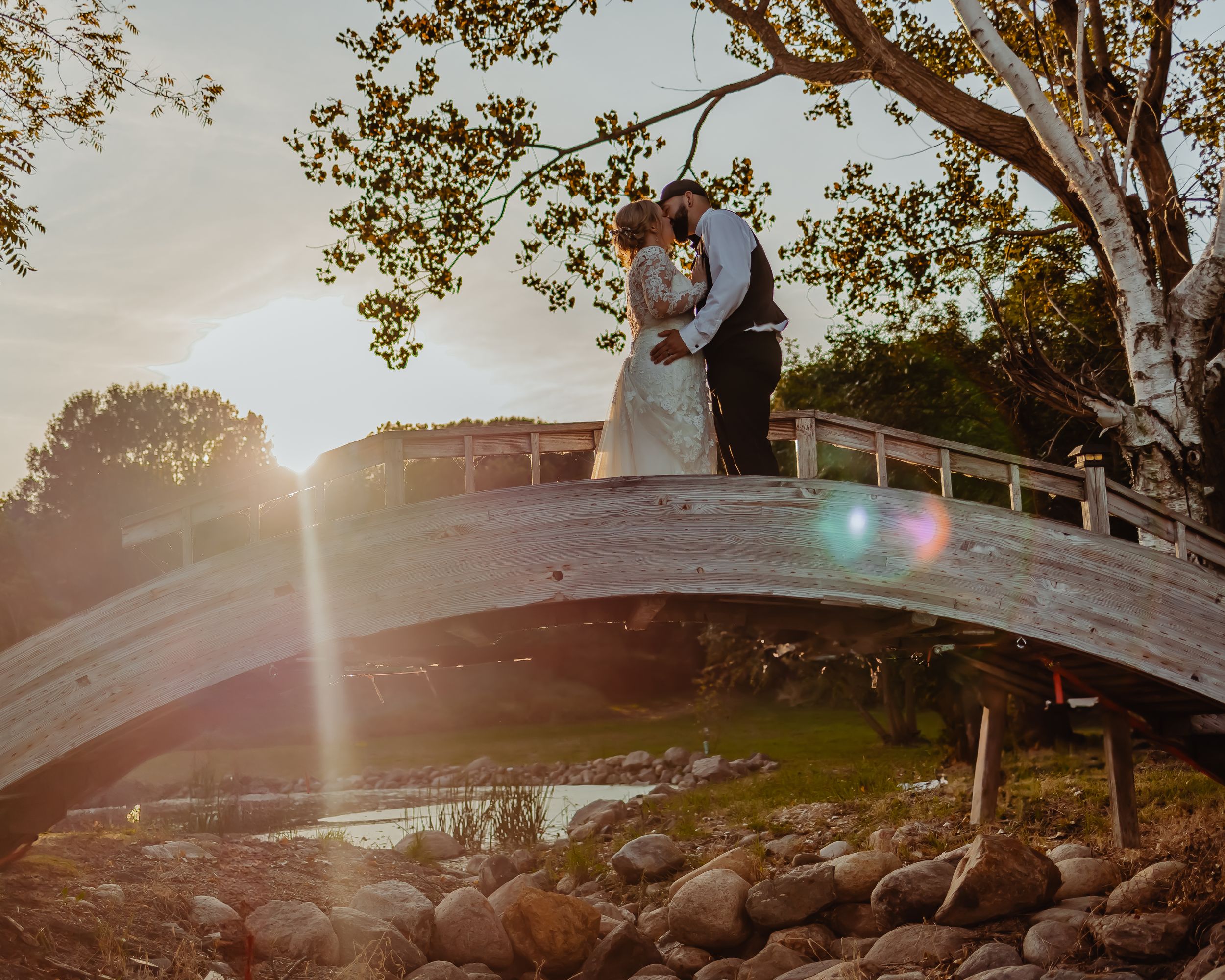 A romantic sunset wedding moment on a curved bridge over a rocky creek with golden sunlight streaming through trees.