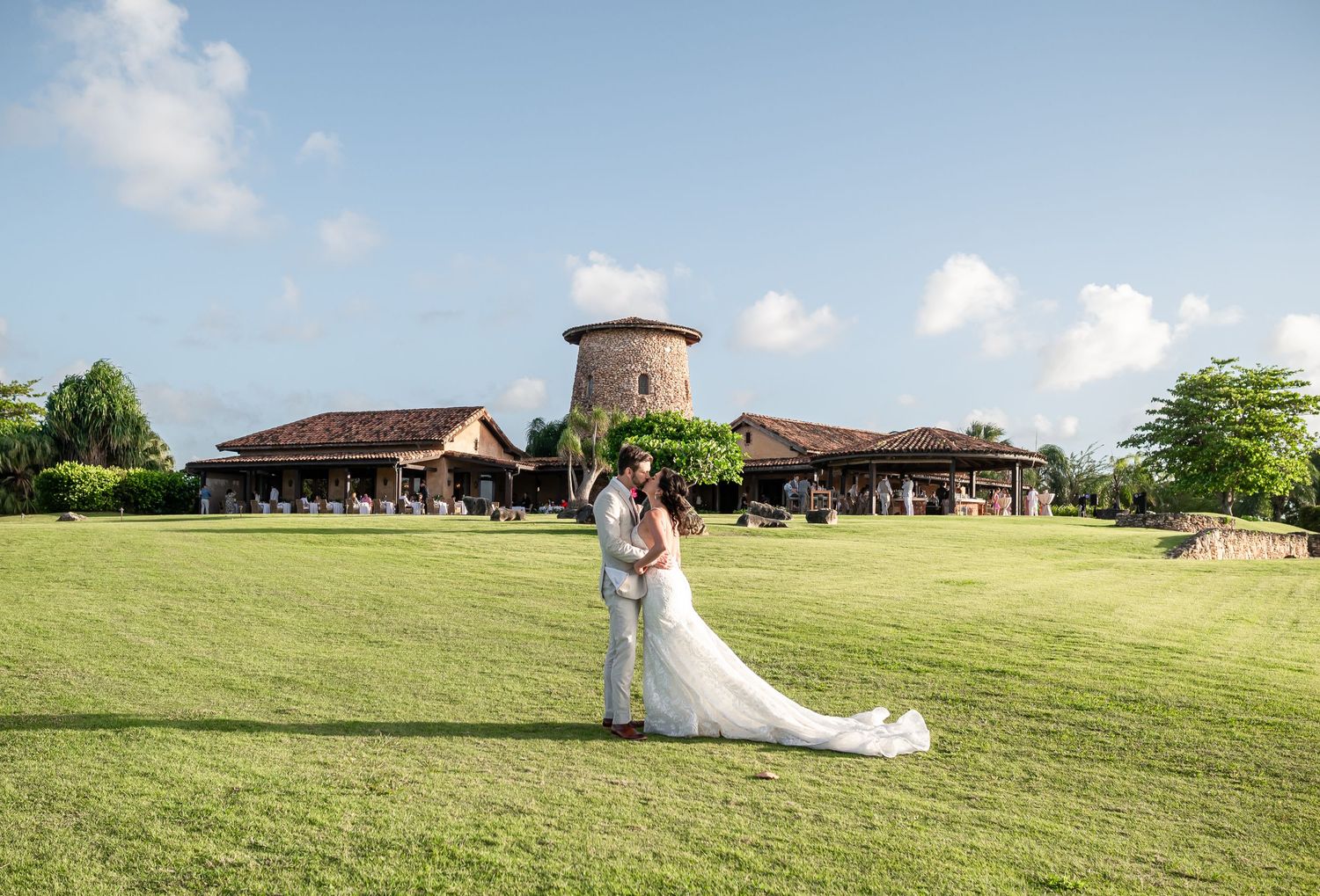 A couple poses on a sunny lawn with a historic tower building in the background.