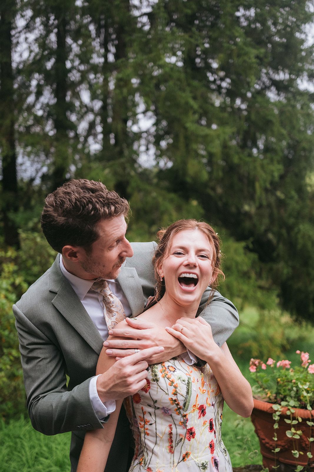A joyful couple share an intimate laughing moment together outdoors with green trees in the background.