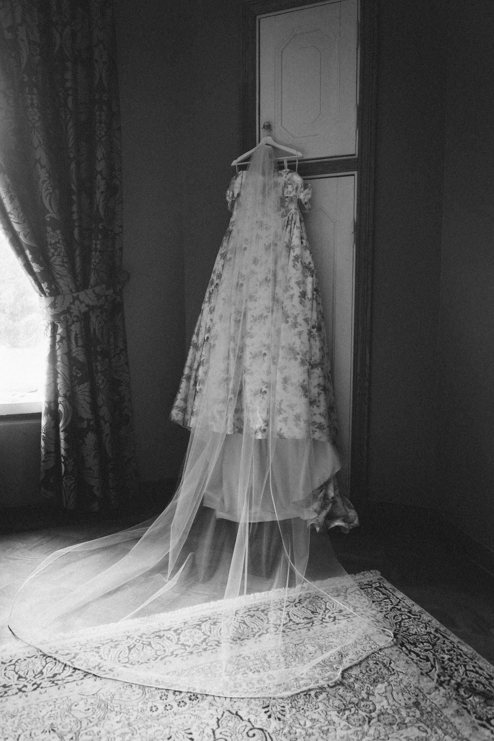 Wedding dress with floral details and long veil hanging on door in elegant room with curtains and decorative rug.