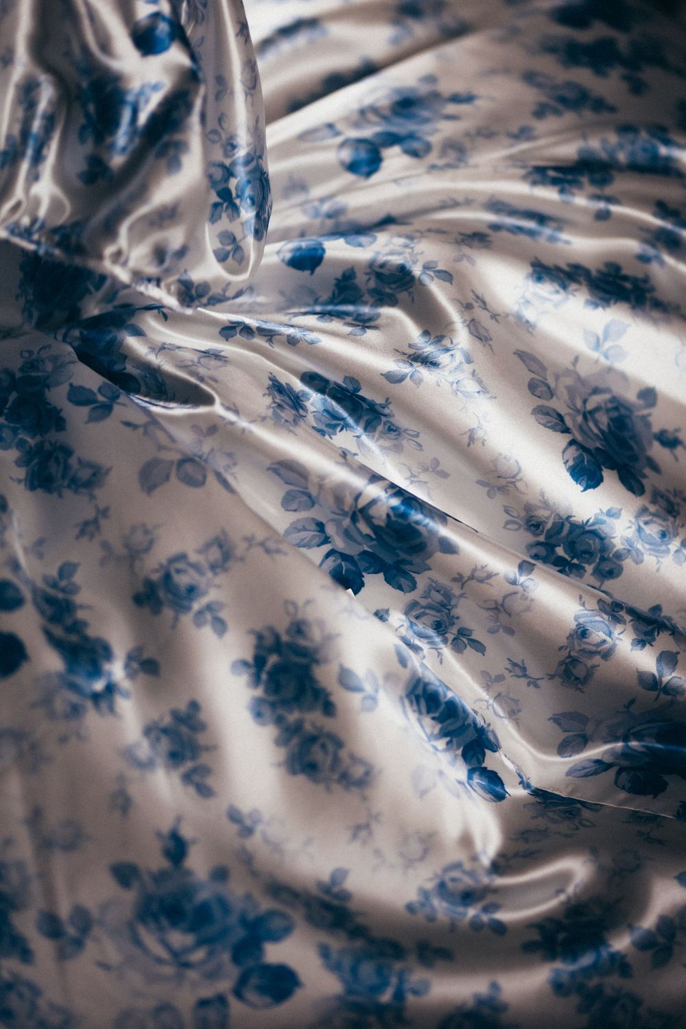 Luxurious satin fabric with blue floral rose print, draped in soft elegant folds, showcasing a delicate vintage-inspired pattern.