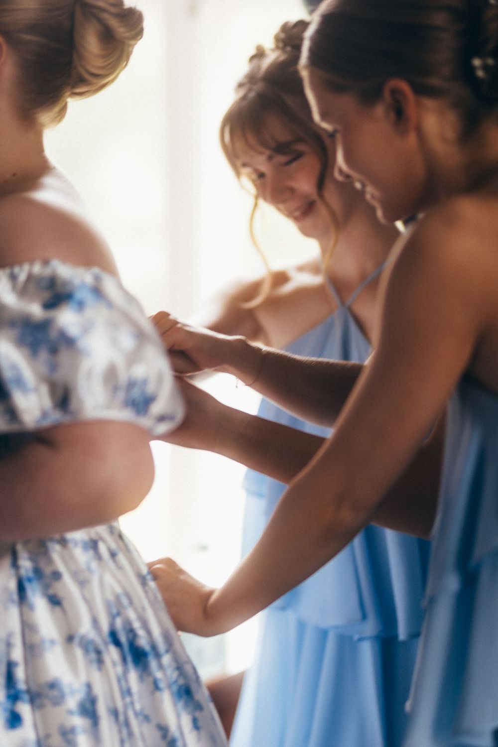 Bridesmaids in light blue dresses helping each other get ready, smiling and adjusting details before a wedding ceremony.