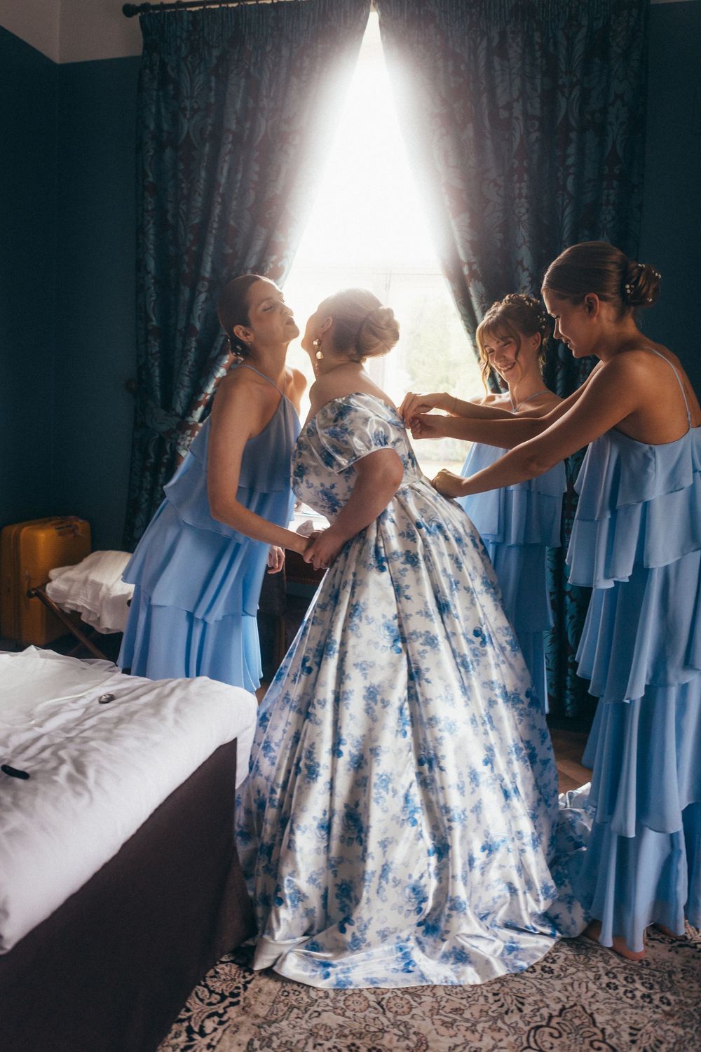 Bridesmaids in blue dresses help bride into floral ballgown by sunlit window in elegant room.