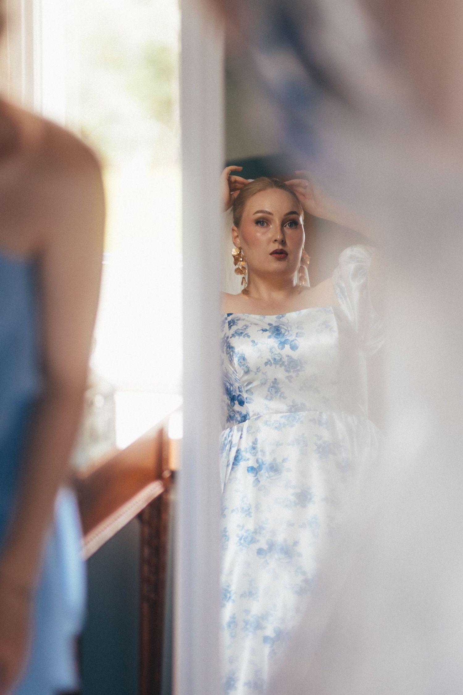 Woman in blue floral dress with gold earrings posing elegantly near a mirror in a bright, airy room.