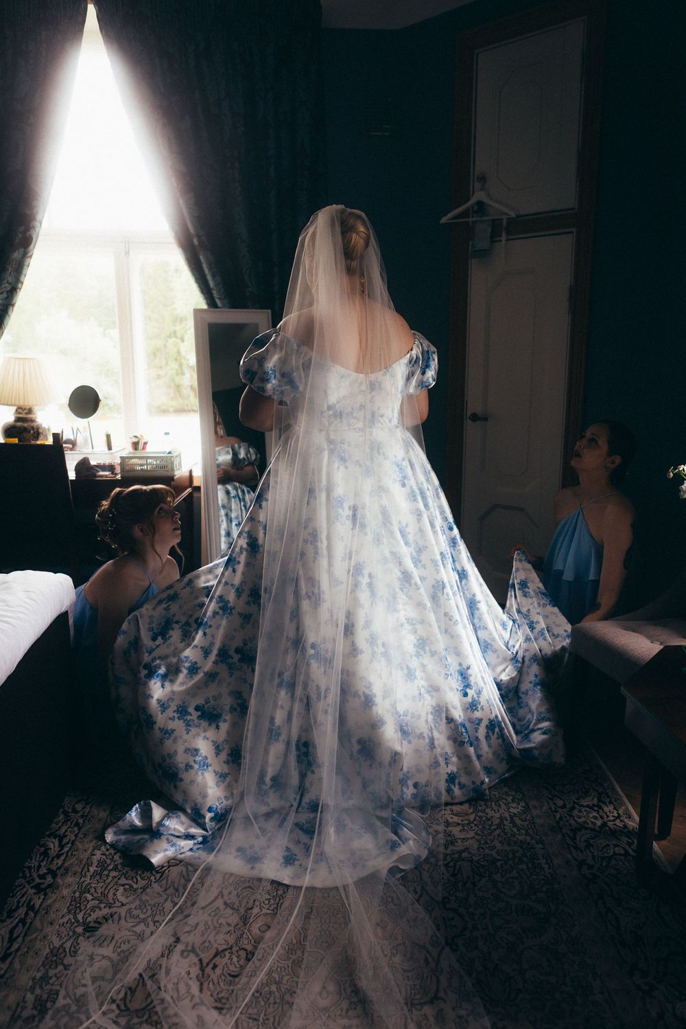 Bride in blue floral wedding gown with veil, assisted by bridesmaids in blue dresses in an elegant dark room.