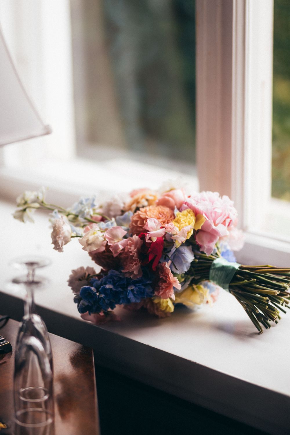 Colorful wedding bouquet with pink, blue, and yellow flowers resting on a white windowsill beside wine glasses.