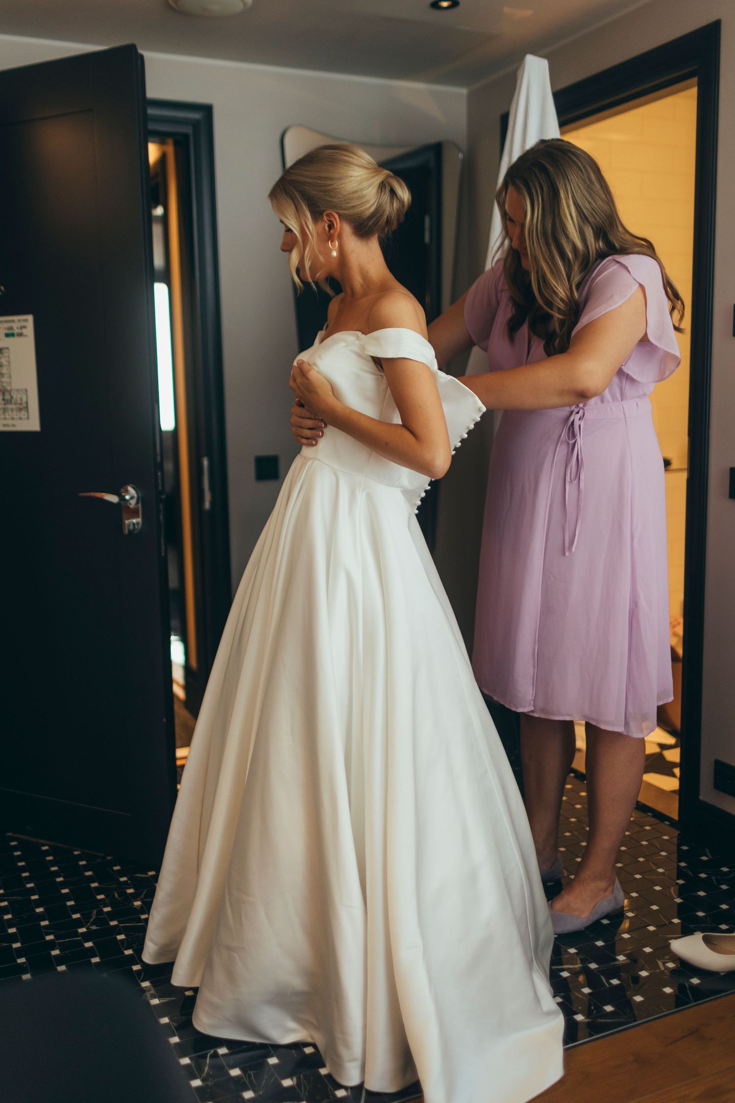 Bridesmaids in lilac dresses helping with wedding dress preparations.