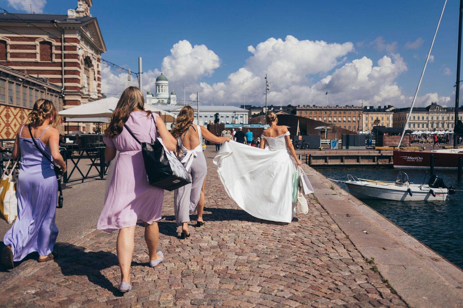 Wedding party walking along a sunny harbor promenade with boats in background.