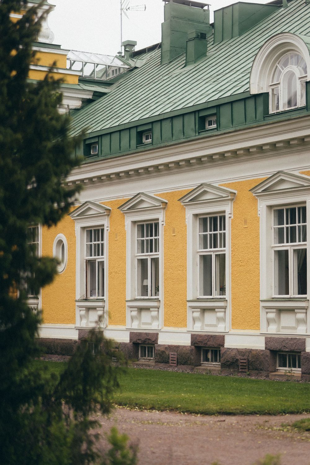 Historic yellow building with white trim, green metal roof, and classical architectural details surrounded by evergreen trees.