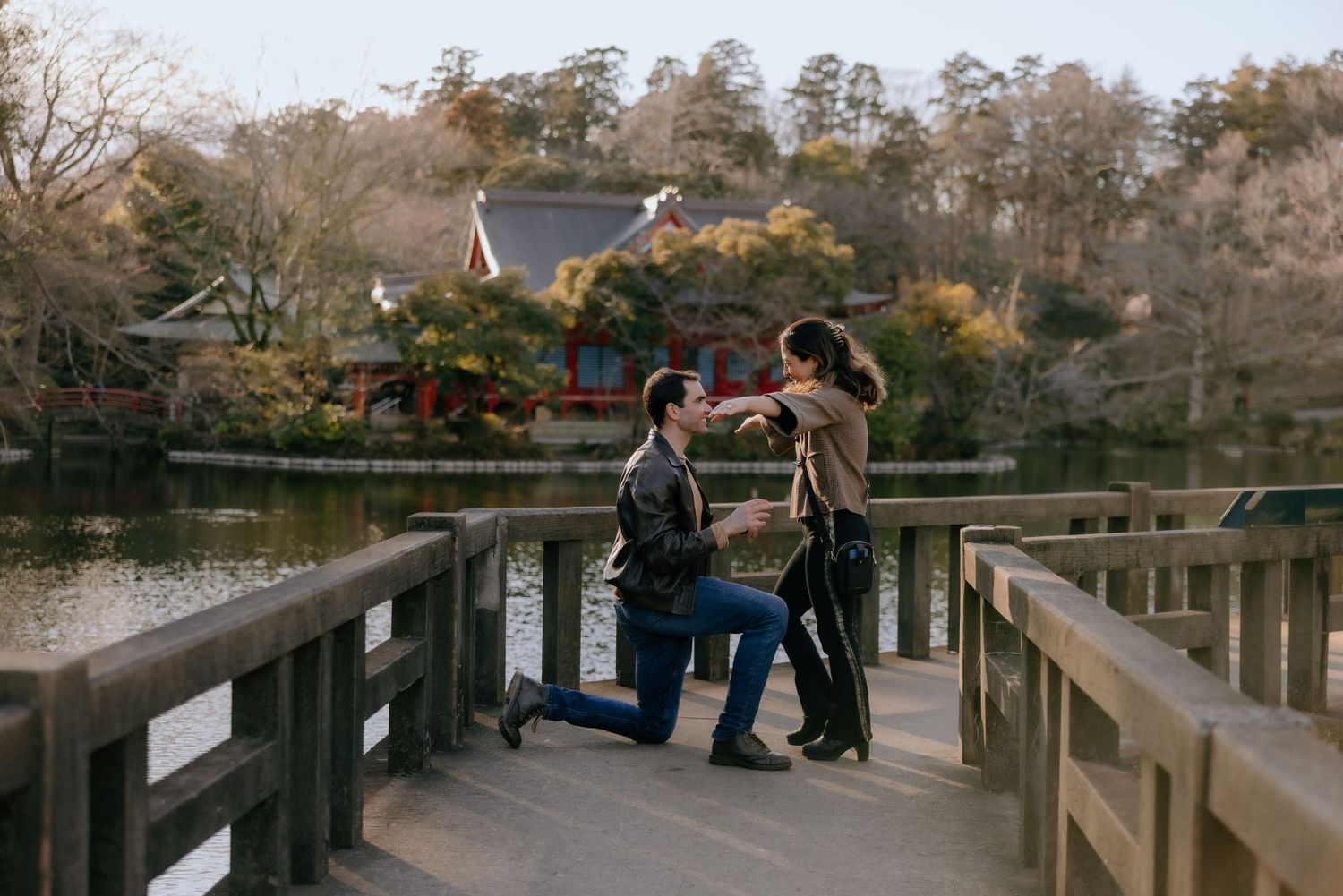 A romantic proposal scene takes place on a wooden bridge over a lake surrounded by autumn trees at sunset.