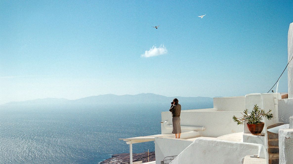 Person standing on a white terrace overlooking the sea and distant mountains on a sunny day.