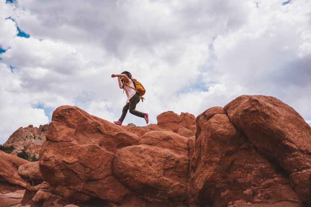 A person jumps between large red rock formations under a cloudy sky.