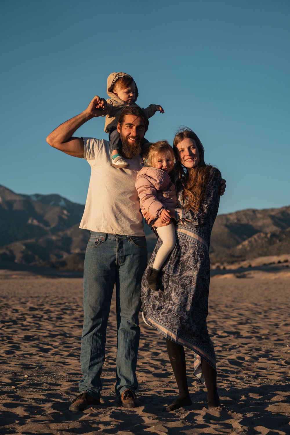 A family shares a tender moment together on sandy terrain with mountains in the background at sunset.