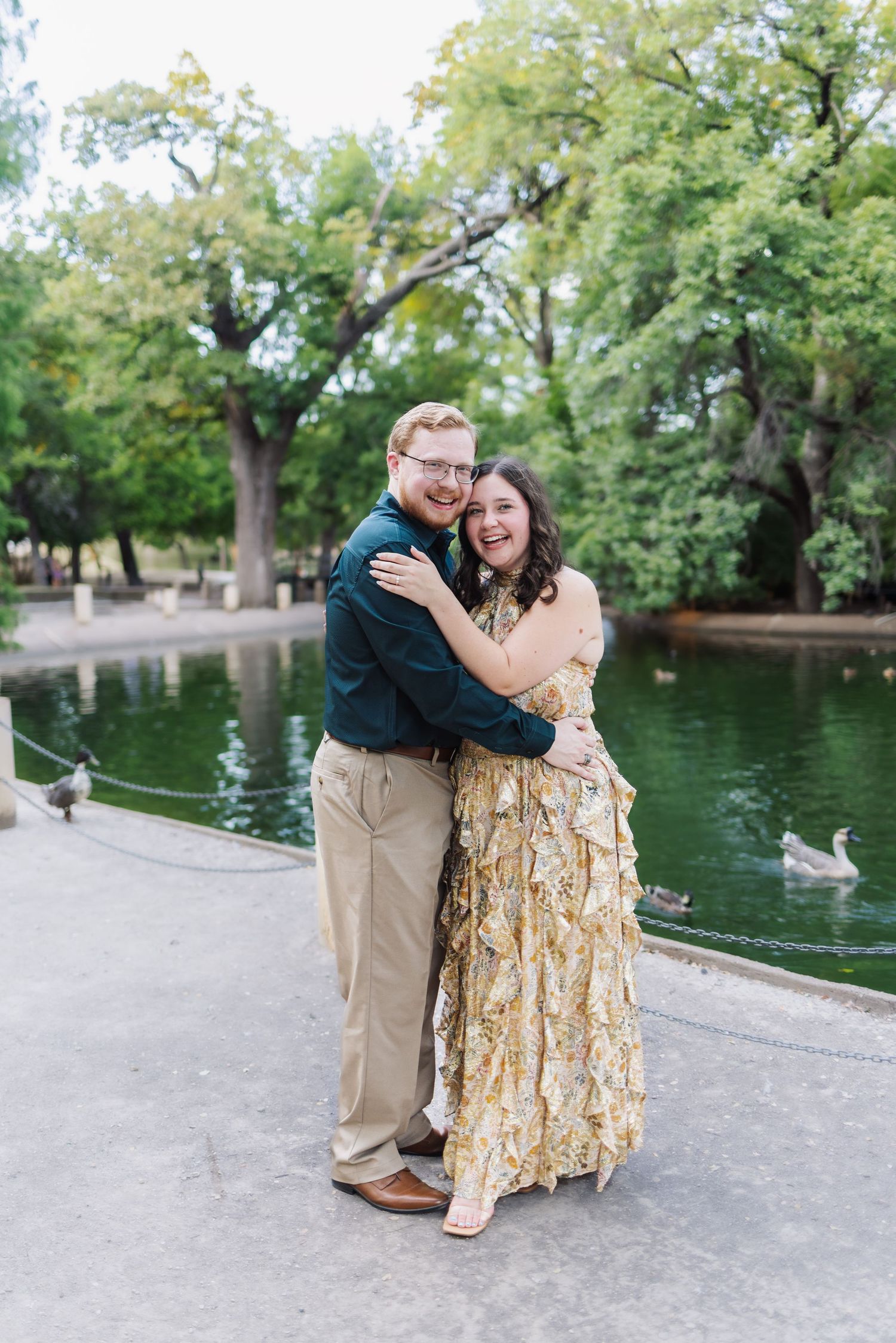 A couple embraces by a pond with ducks swimming in the background on a sunny day in a park setting.