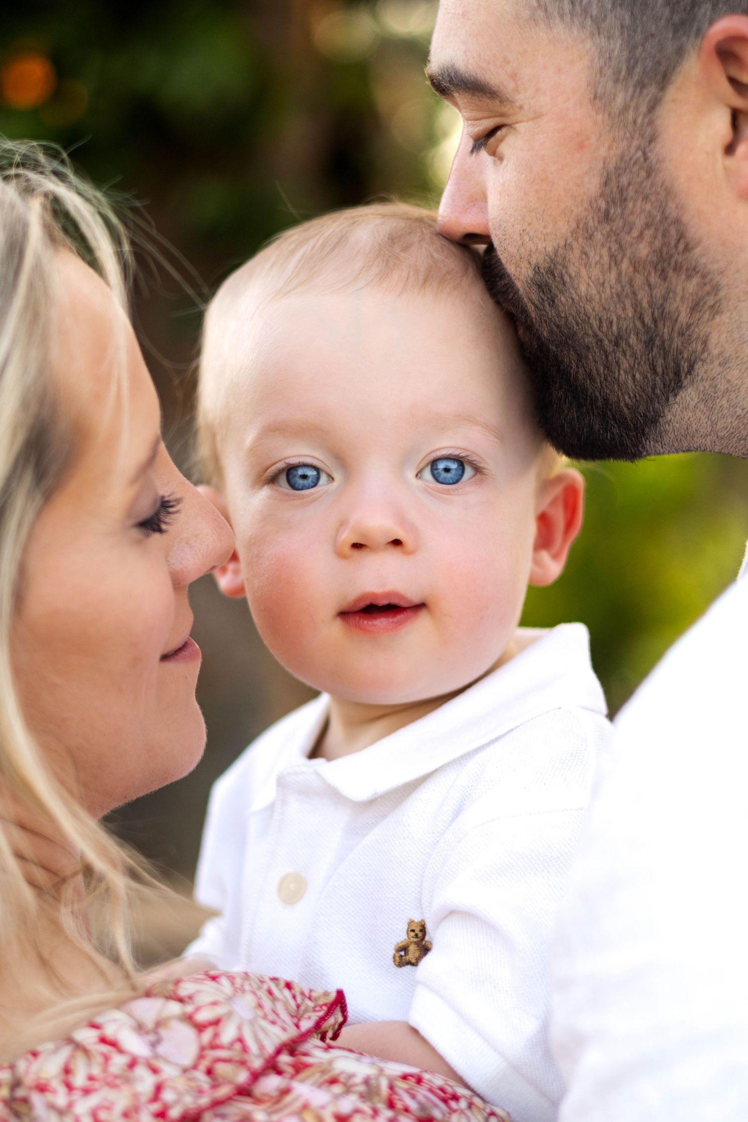 A baby with bright blue eyes wearing a white polo shirt is embraced in a tender family moment outdoors.