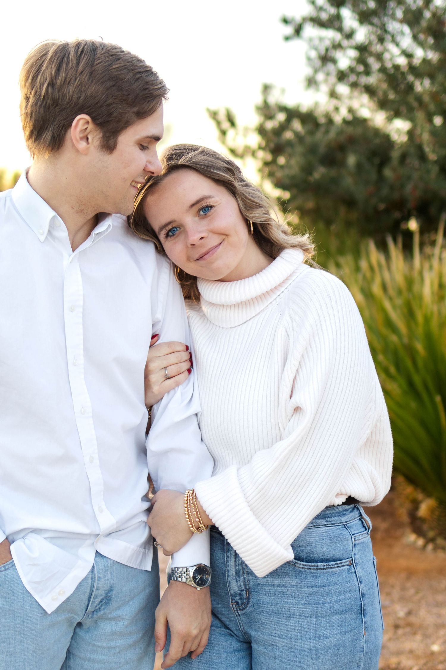 A couple embraces outdoors, with one wearing a white sweater and jeans, and the other in a white button-down shirt.
