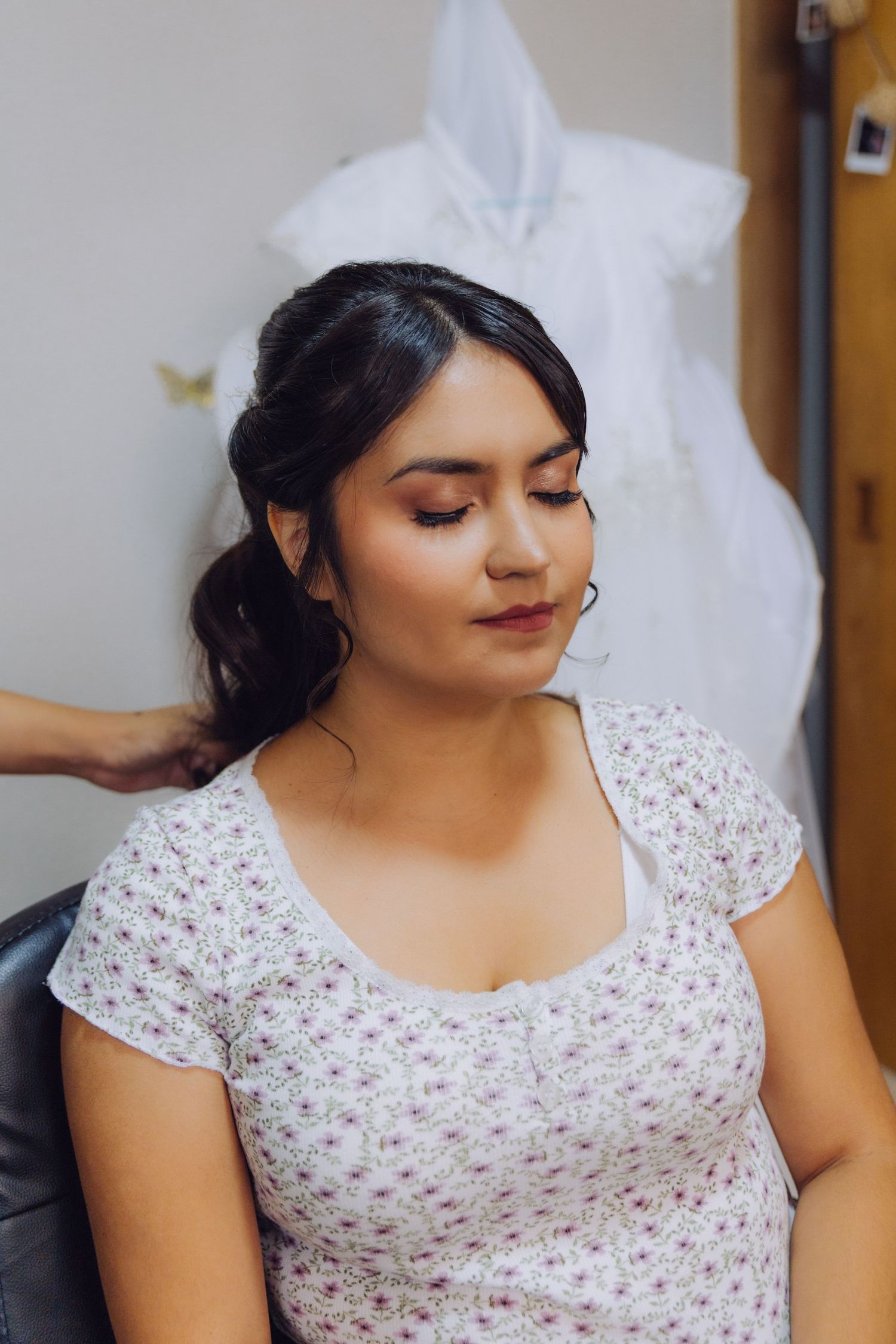 Bride getting hair and makeup done before wedding with white gown hanging in background.