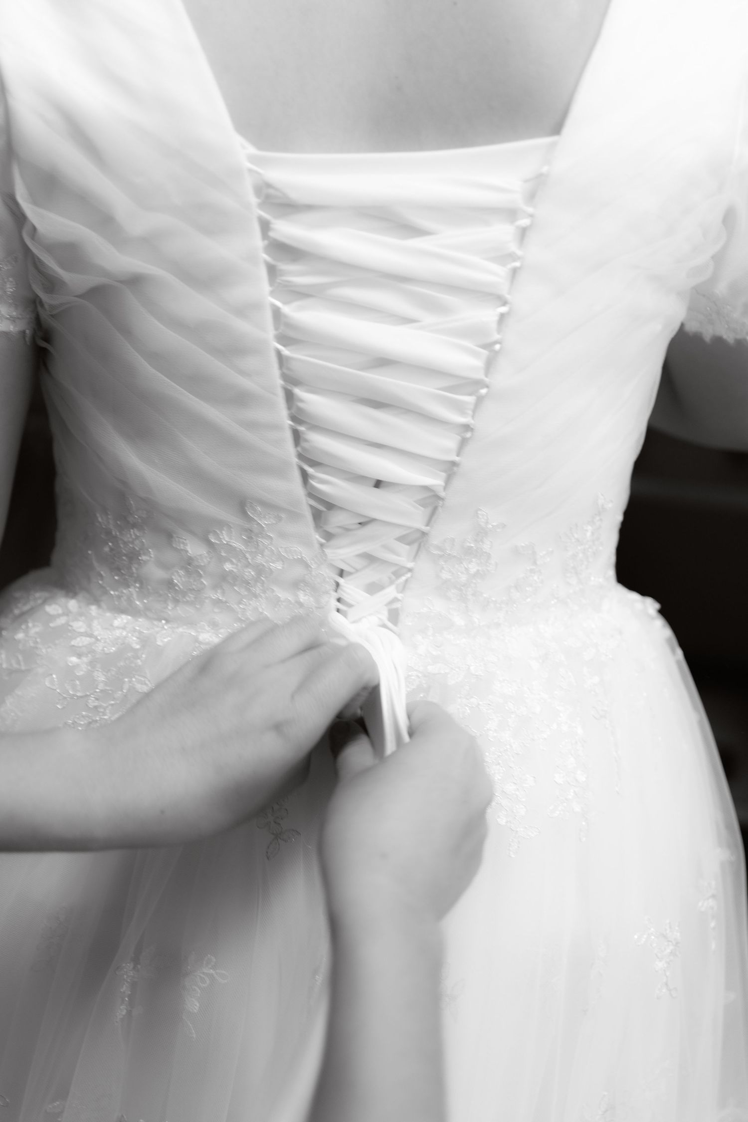 Black and white close-up of wedding dress corset back being laced up by bridesmaid.