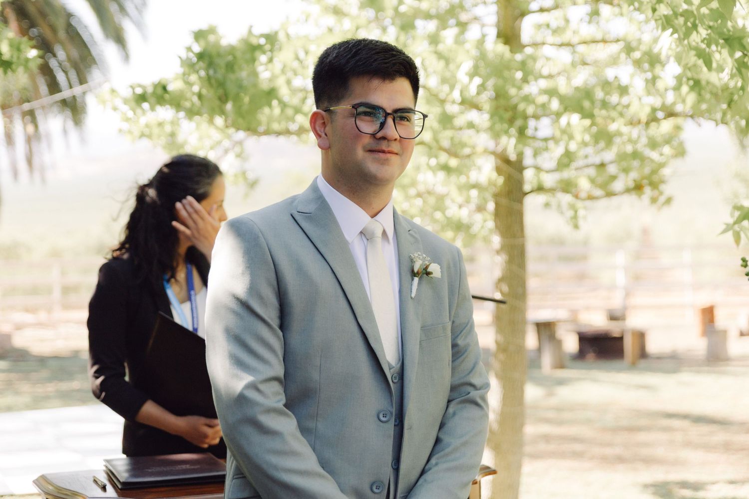 Groom in light gray suit smiling warmly while waiting at the wedding ceremony altar.