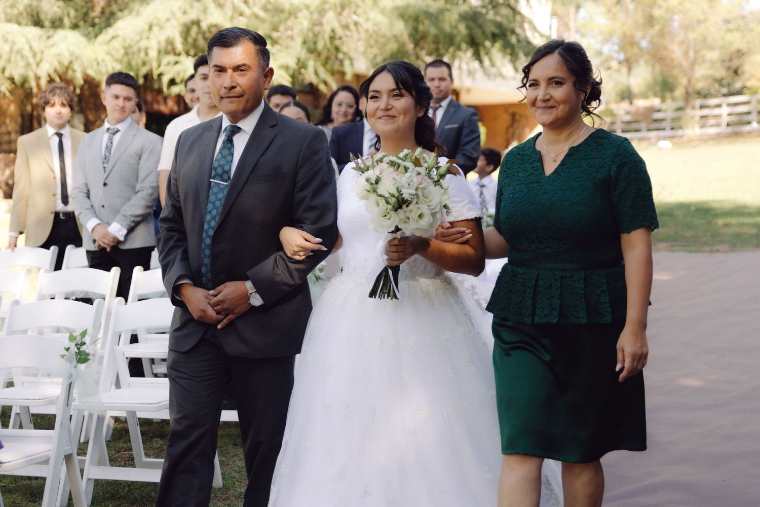 Bride walking down the aisle escorted by her parents at an outdoor wedding ceremony.