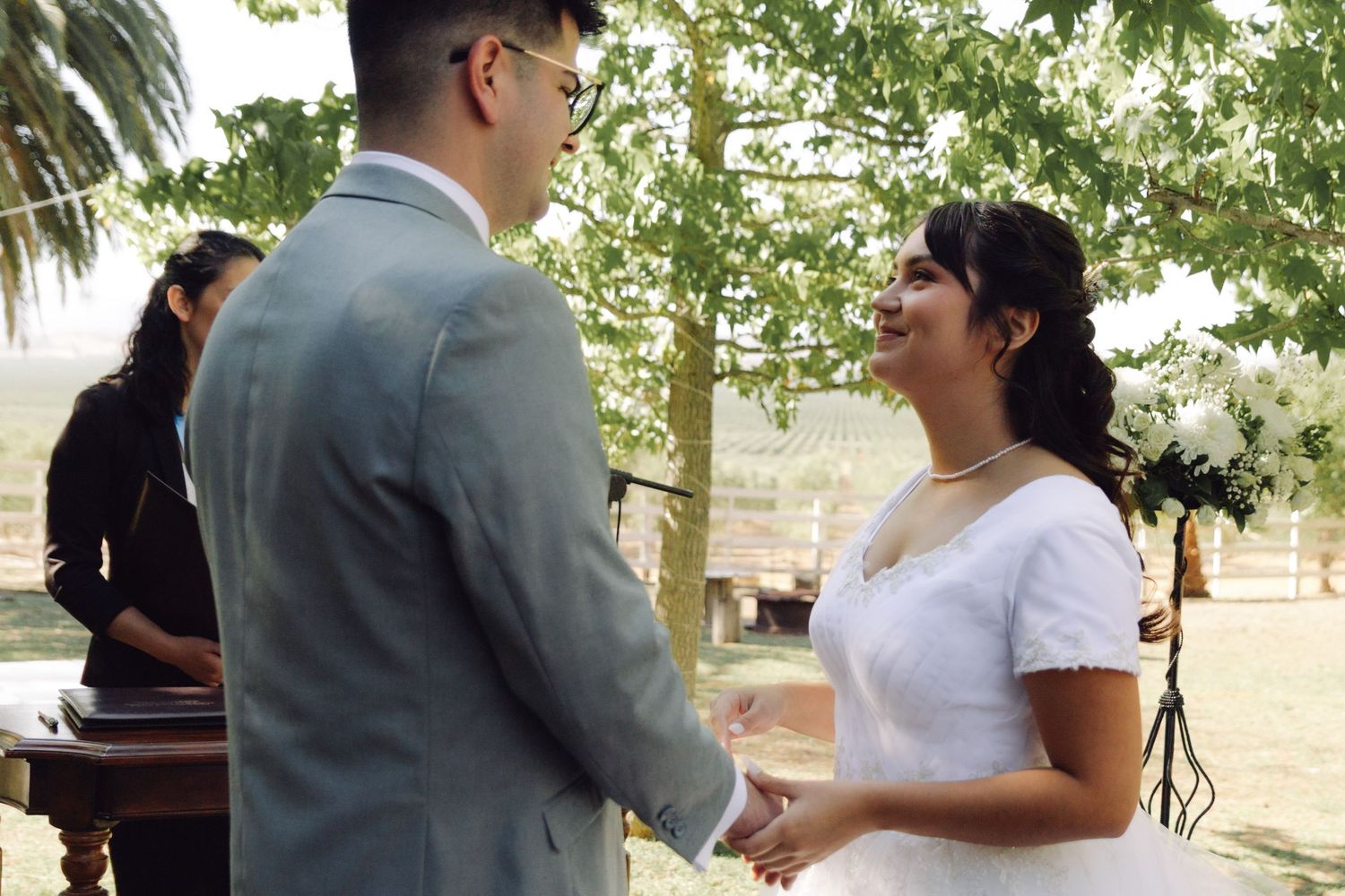 Bride smiling joyfully while holding hands with groom during outdoor wedding ceremony.
