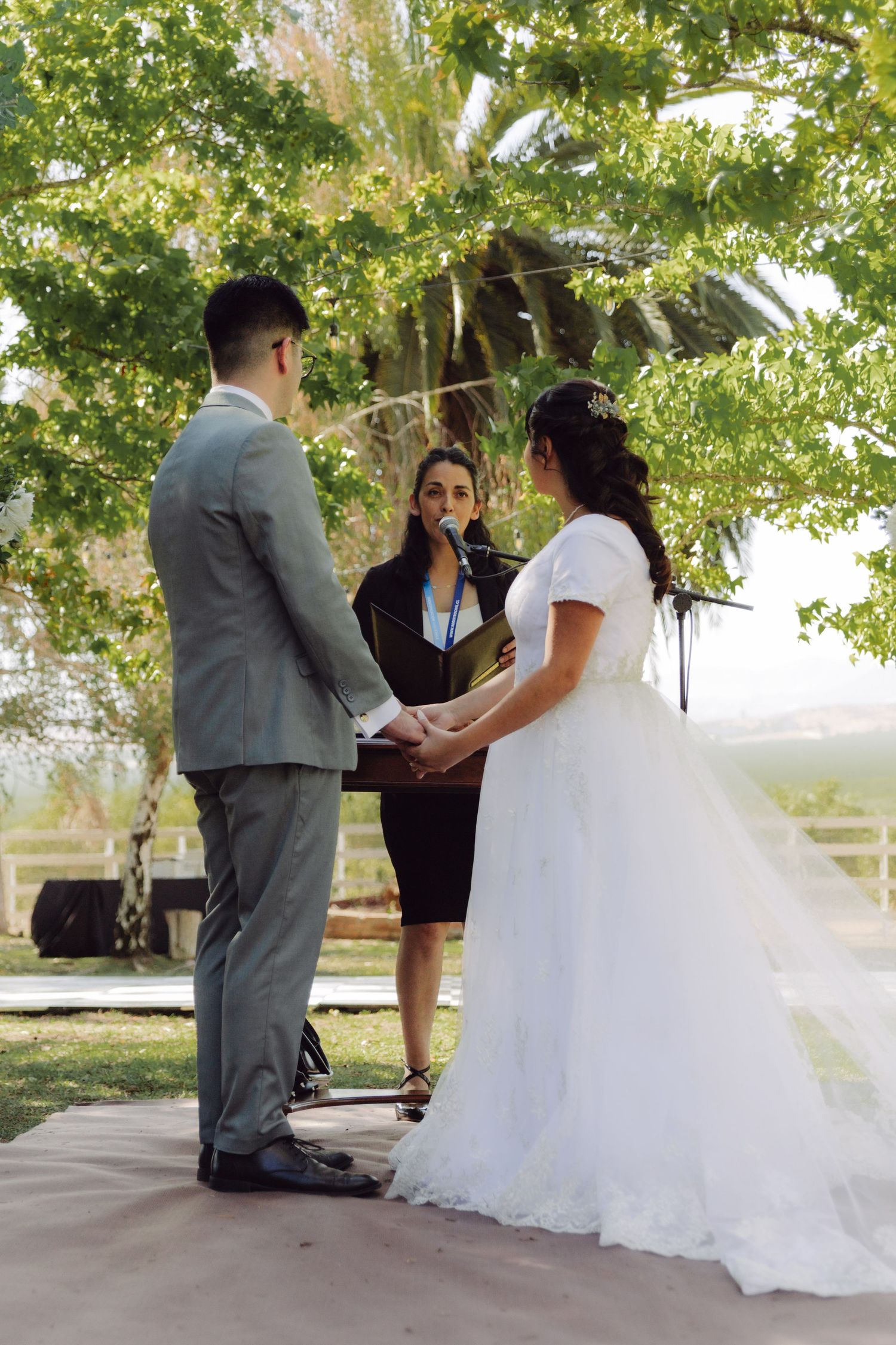 Bride and groom hold hands during outdoor wedding ceremony while officiant speaks at a microphone.