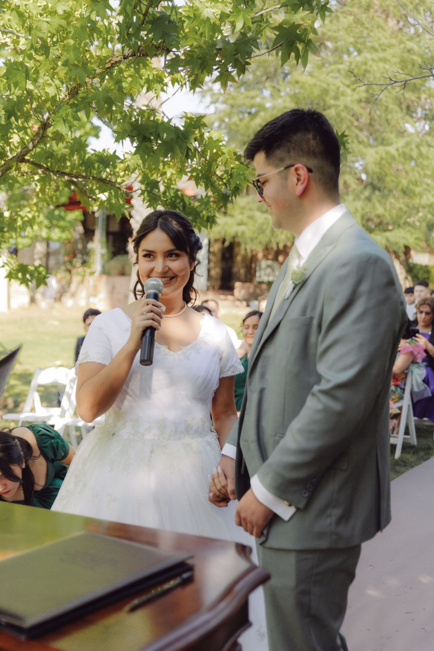 Smiling bride speaks into microphone during outdoor wedding ceremony while groom watches lovingly.