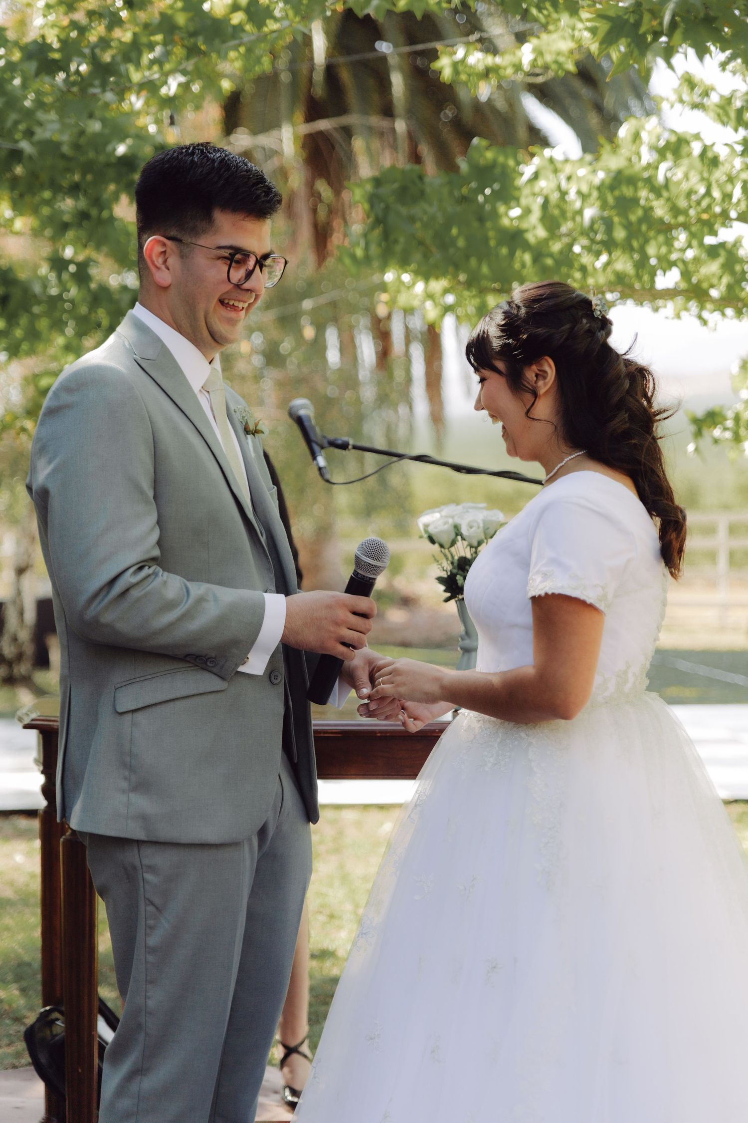 Groom places wedding ring on brides finger during outdoor ceremony while sharing a smile.