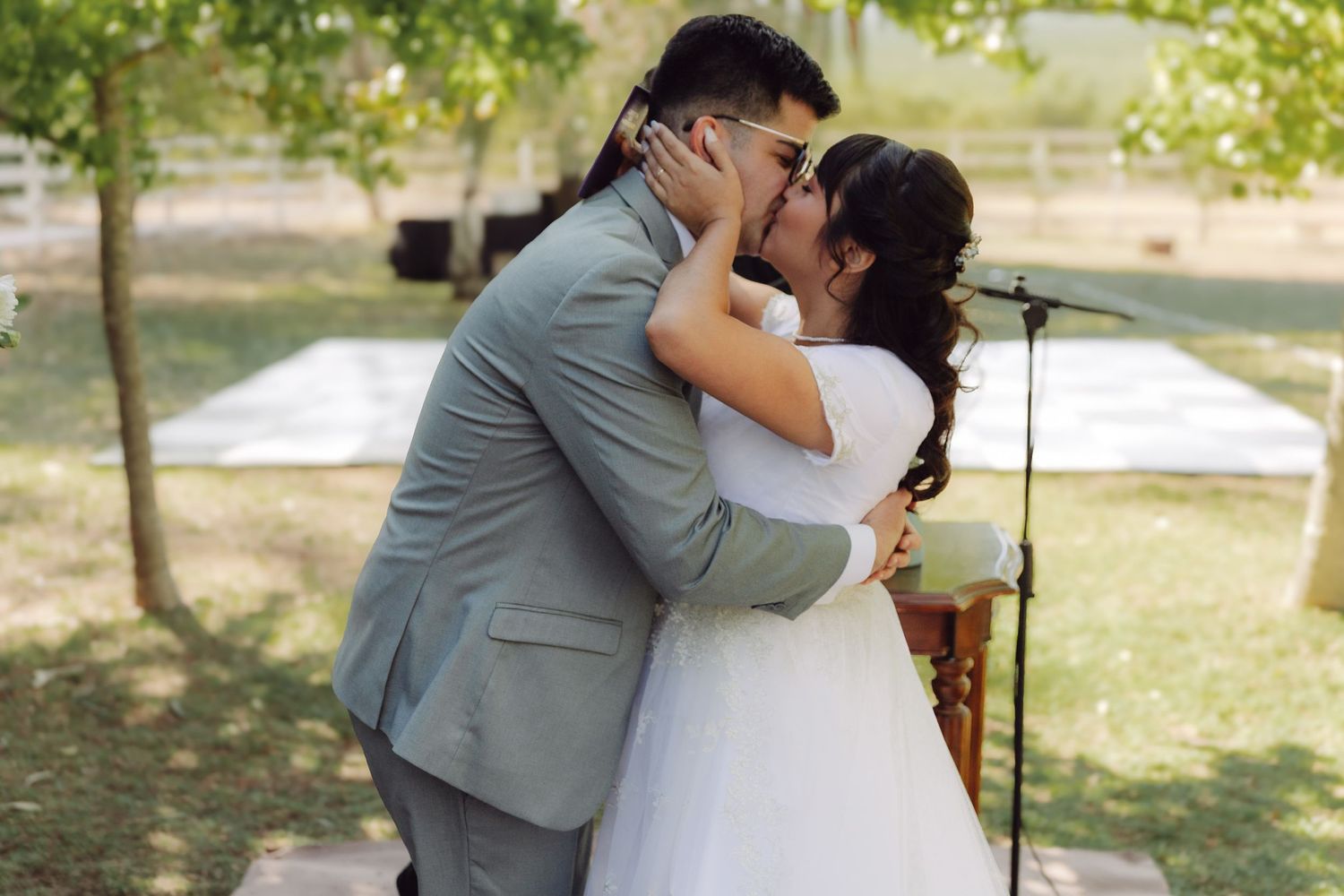 Bride and groom share their first kiss at an outdoor wedding ceremony surrounded by lush green trees.