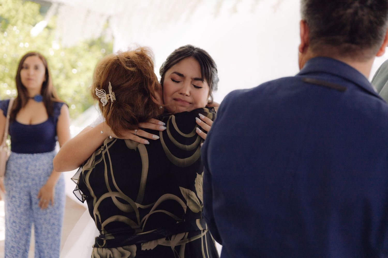 Emotional bride hugs a guest in black floral dress at wedding reception outdoors.
