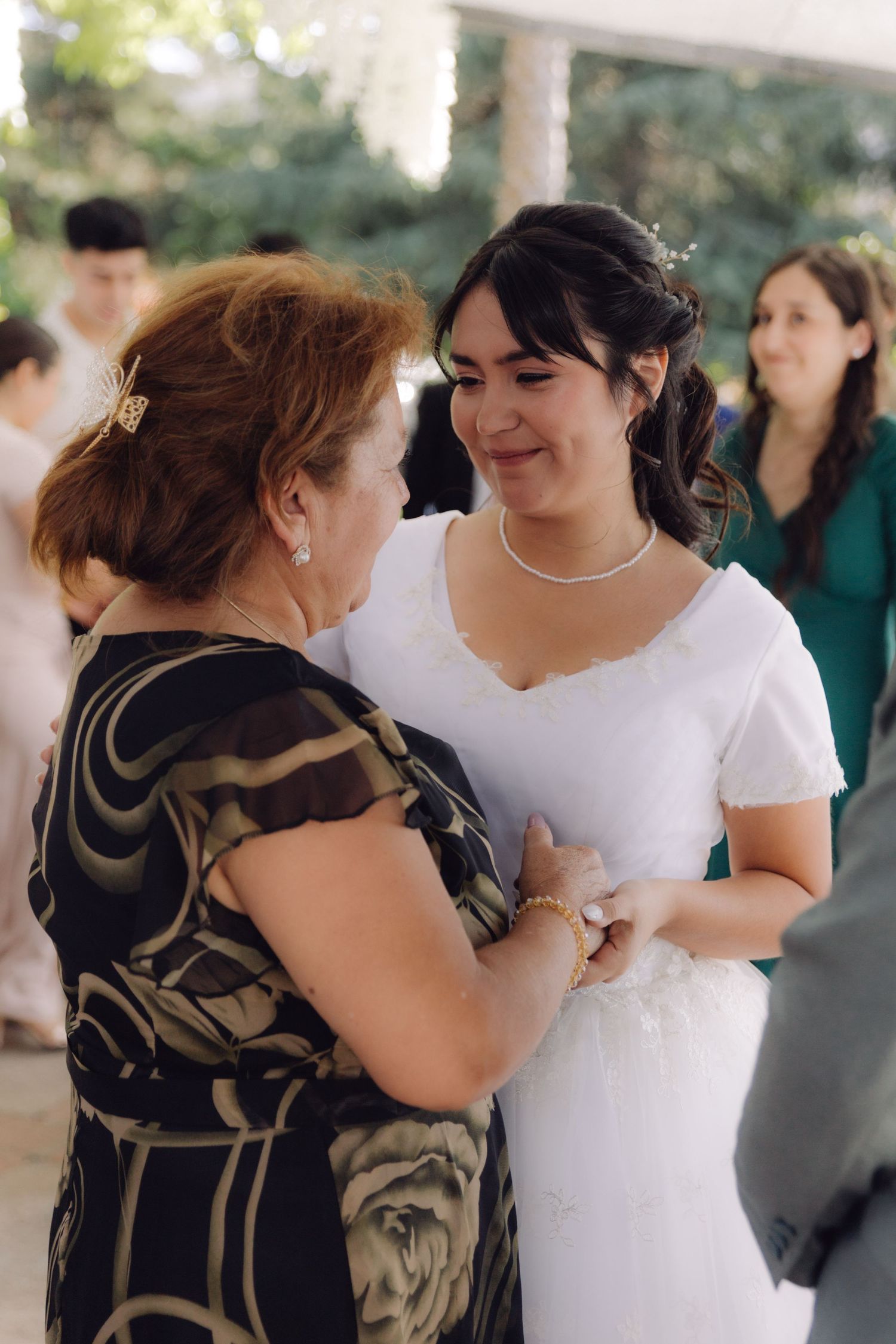 Bride in white dress smiles warmly while holding hands with older woman at wedding.