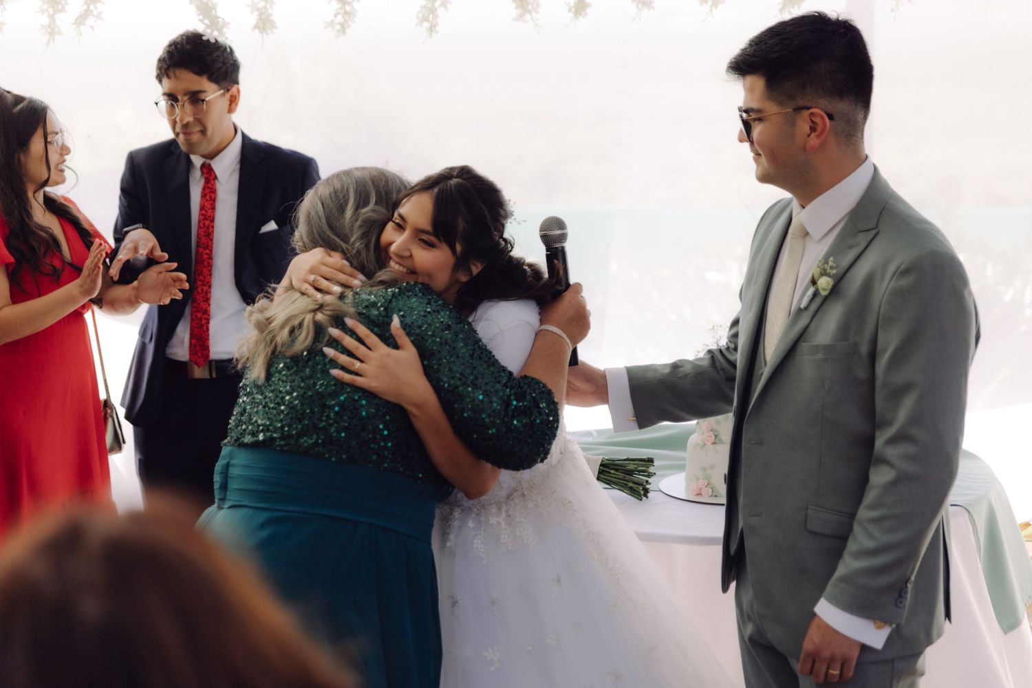 Bride hugs woman in green dress at wedding reception while groom smiles nearby.