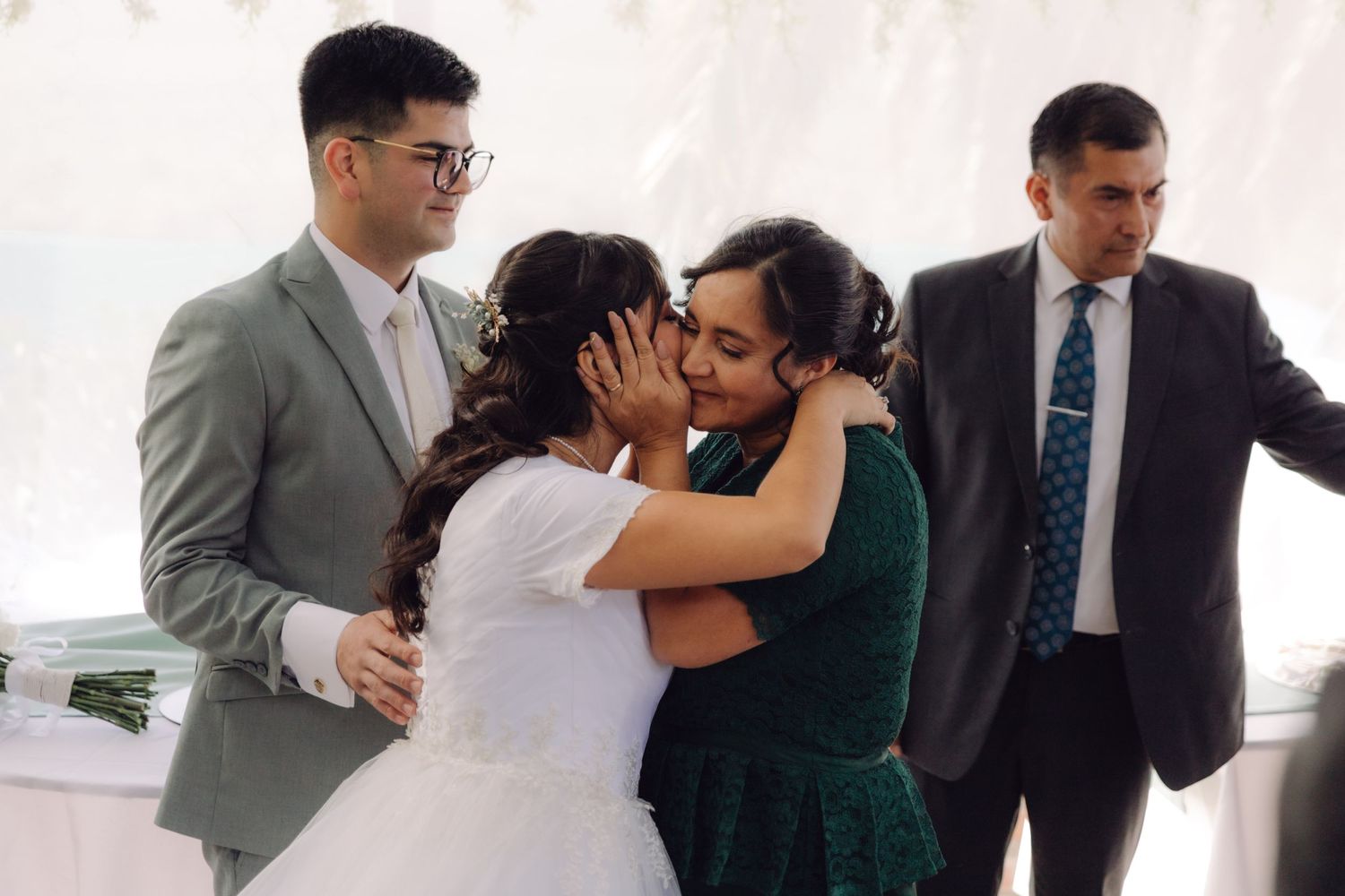 Bride and groom share emotional hugs with guests during wedding ceremony under white tent.