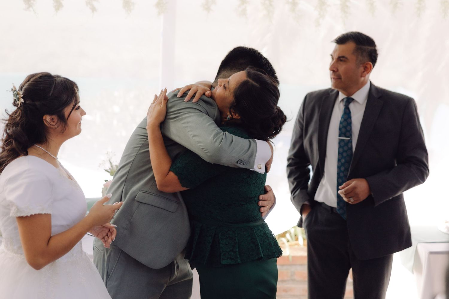 Groom embraces wedding guest warmly while bride and older man look on smiling.