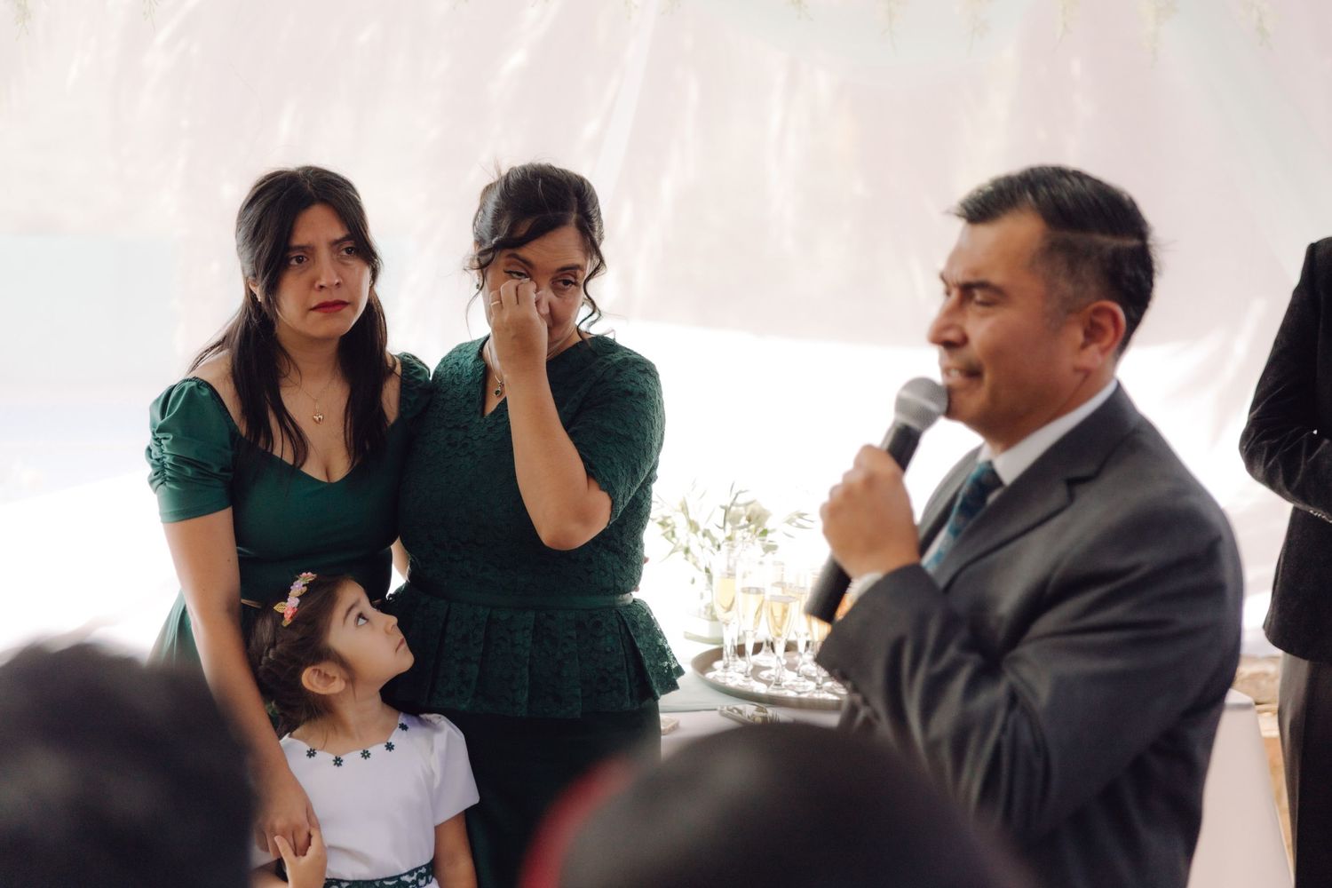 Man speaks into microphone at wedding reception while emotional women and child stand nearby.