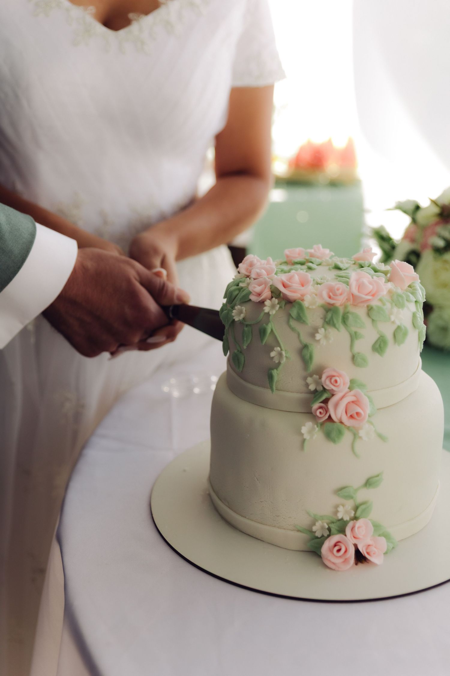 Bride and groom cut a elegant two-tier white wedding cake decorated with pink roses and green leaves.