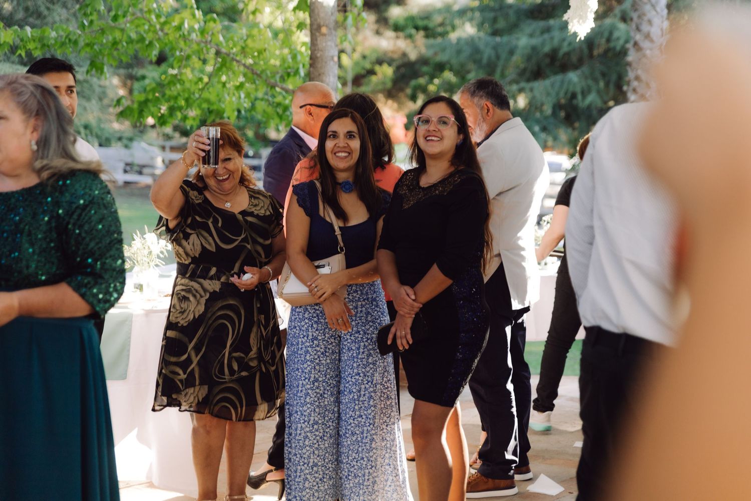 Guests smile and mingle outdoors at a sunny wedding reception celebration.
