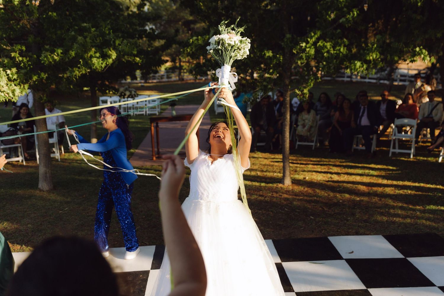 Bride tosses bouquet into the air on checkered dance floor at outdoor wedding.