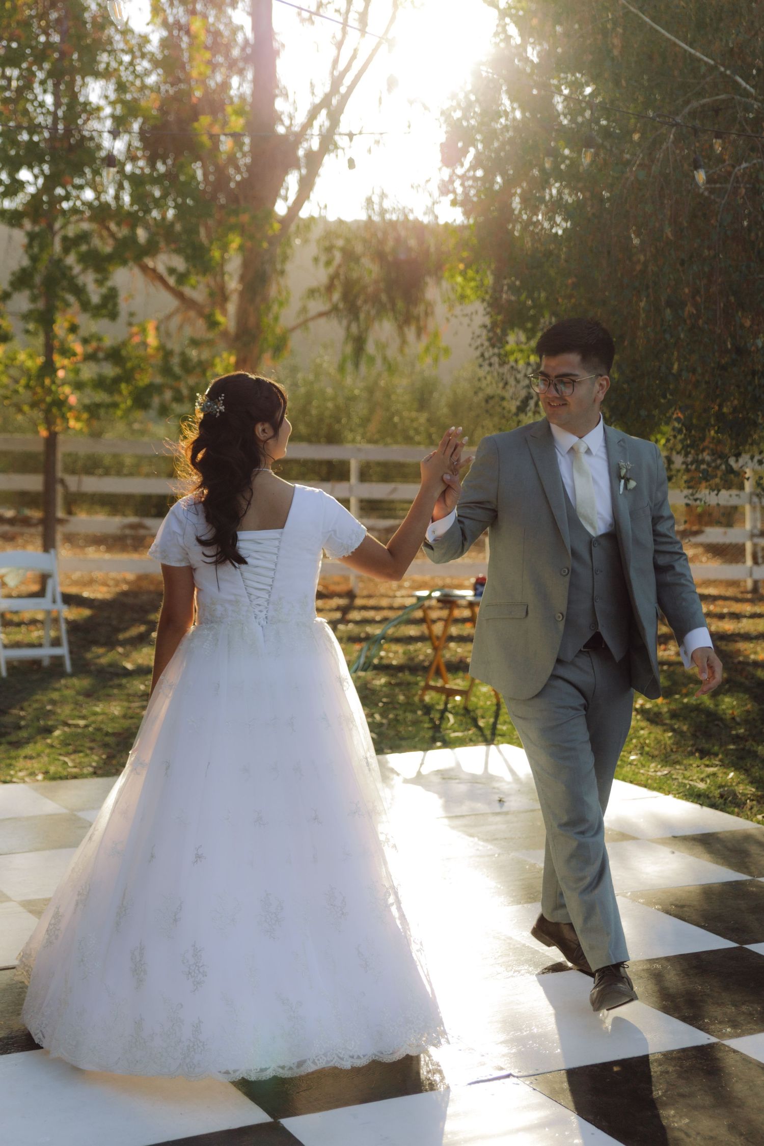 Bride and groom hold hands on a checkered dance floor during their golden hour outdoor reception.