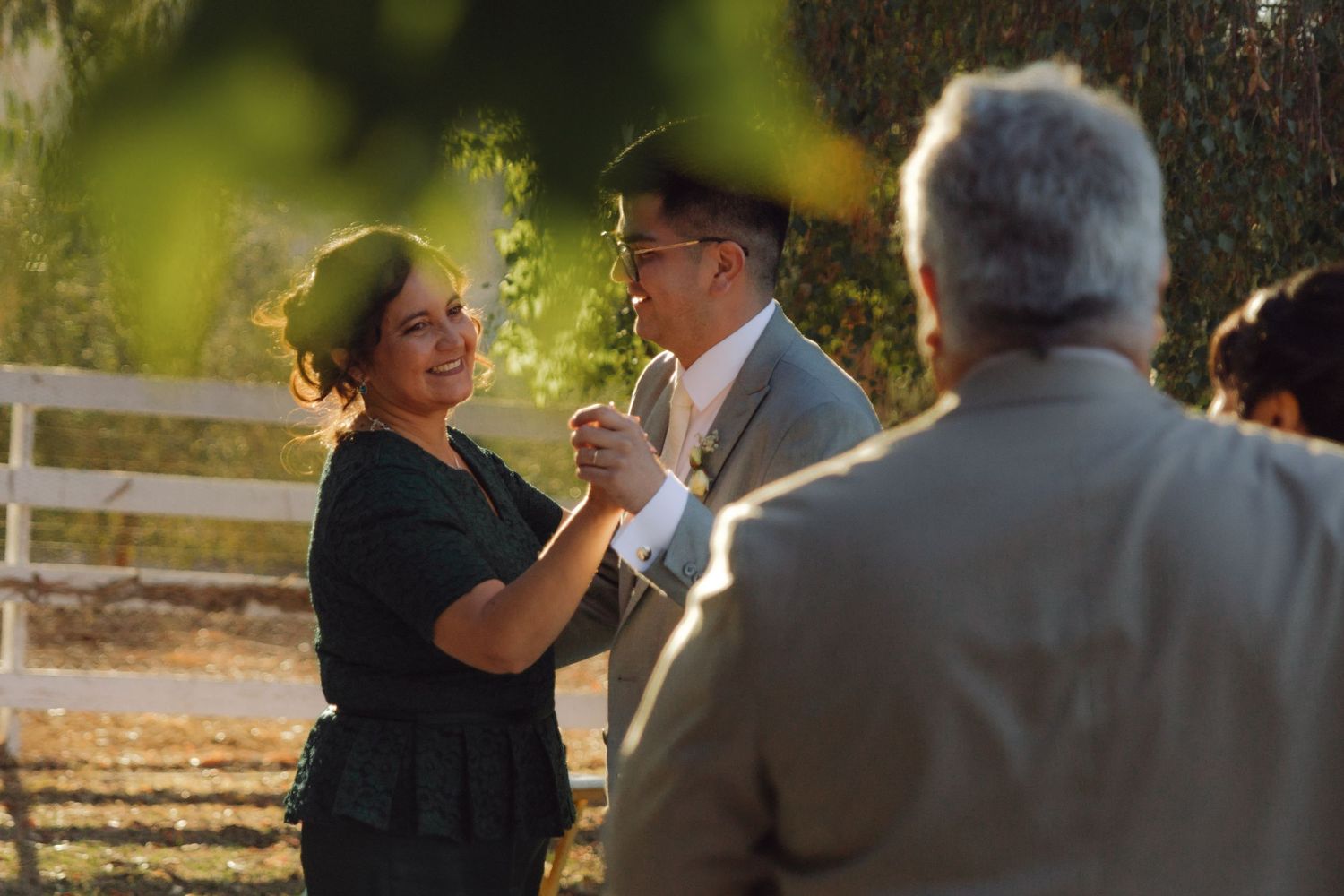 Woman dances with groom outdoors at golden hour wedding reception near white fence.