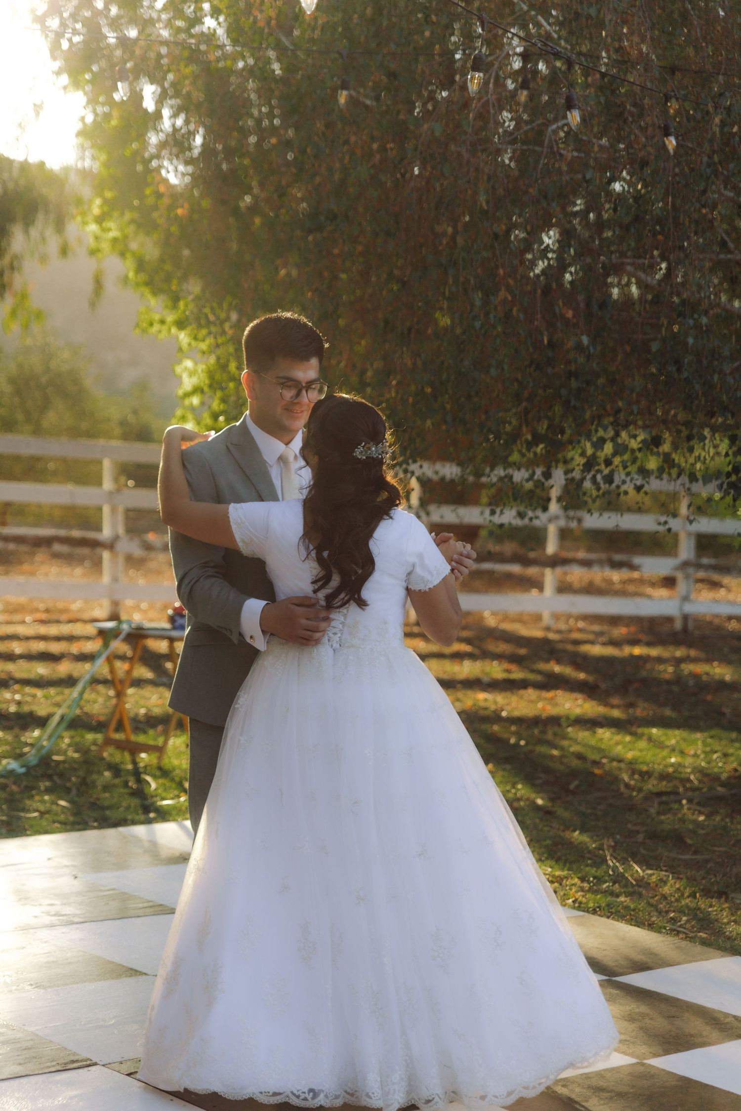 Bride and groom share first dance on checkered floor surrounded by golden sunset light.