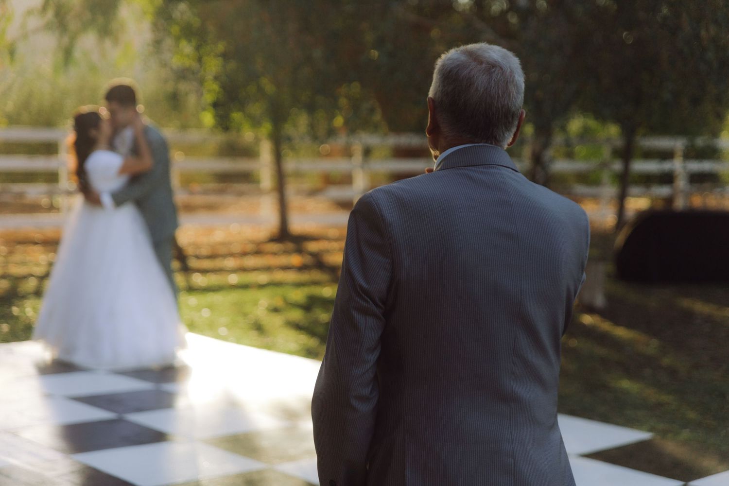 Wedding guest watches as newlyweds share a romantic kiss on a checkered outdoor dance floor.