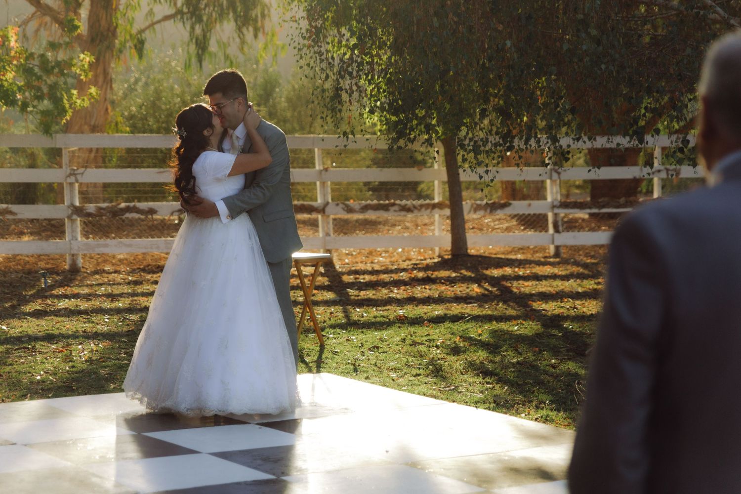 Bride and groom share their first kiss on a checkered dance floor outdoors at a sunny wedding venue.
