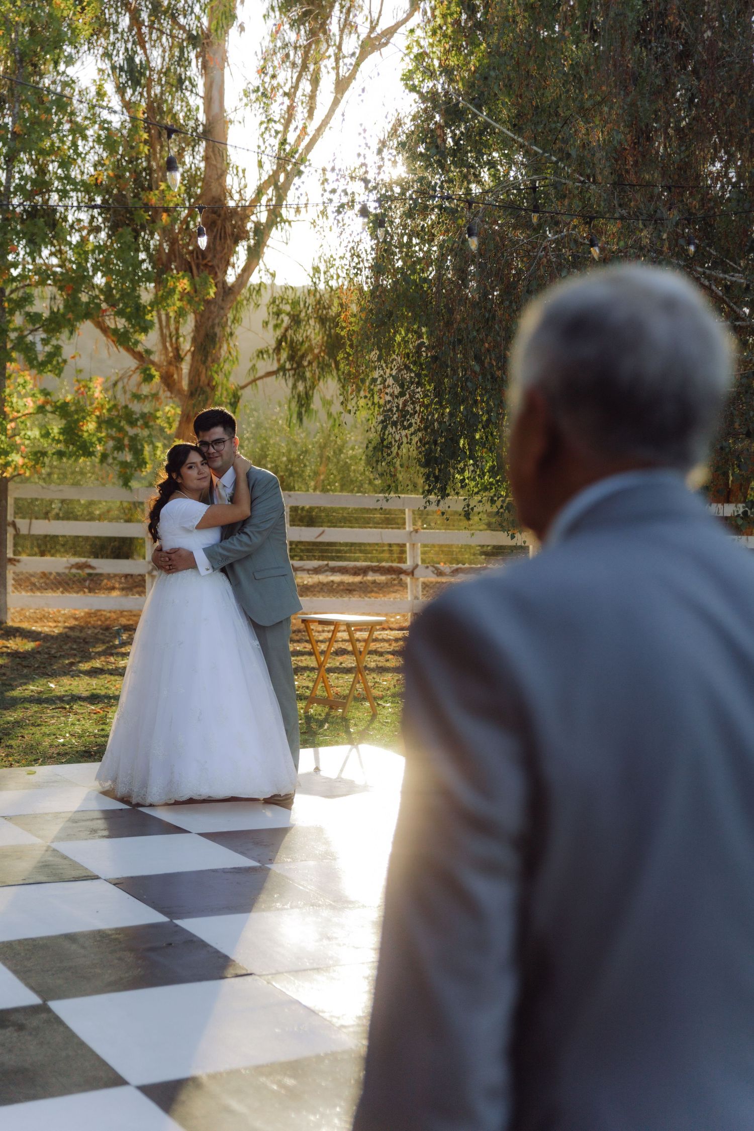 Newlyweds embrace on an outdoor checkered dance floor while a guest watches during wedding reception.