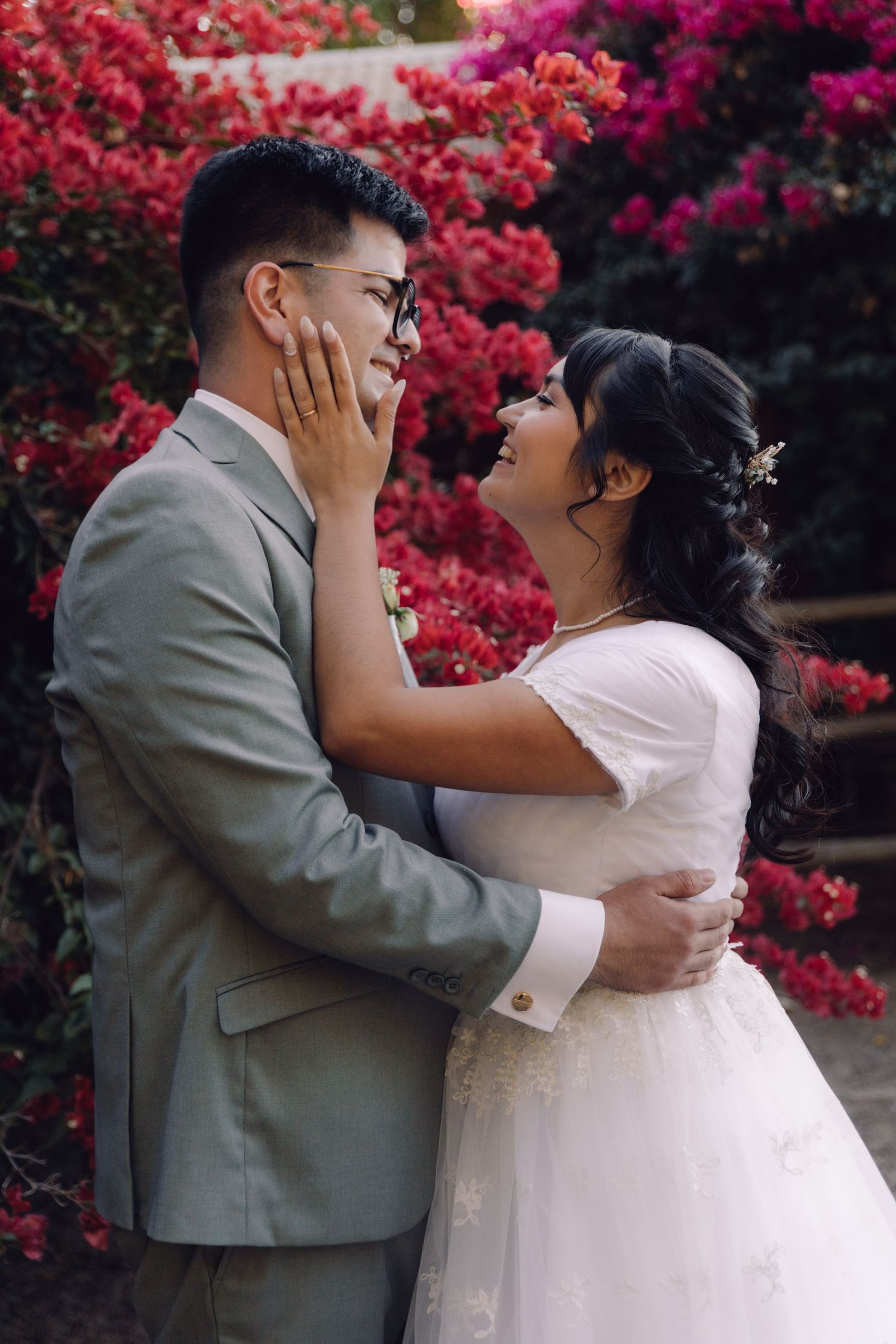 Bride and groom share a romantic moment in front of vibrant red bougainvillea flowers.