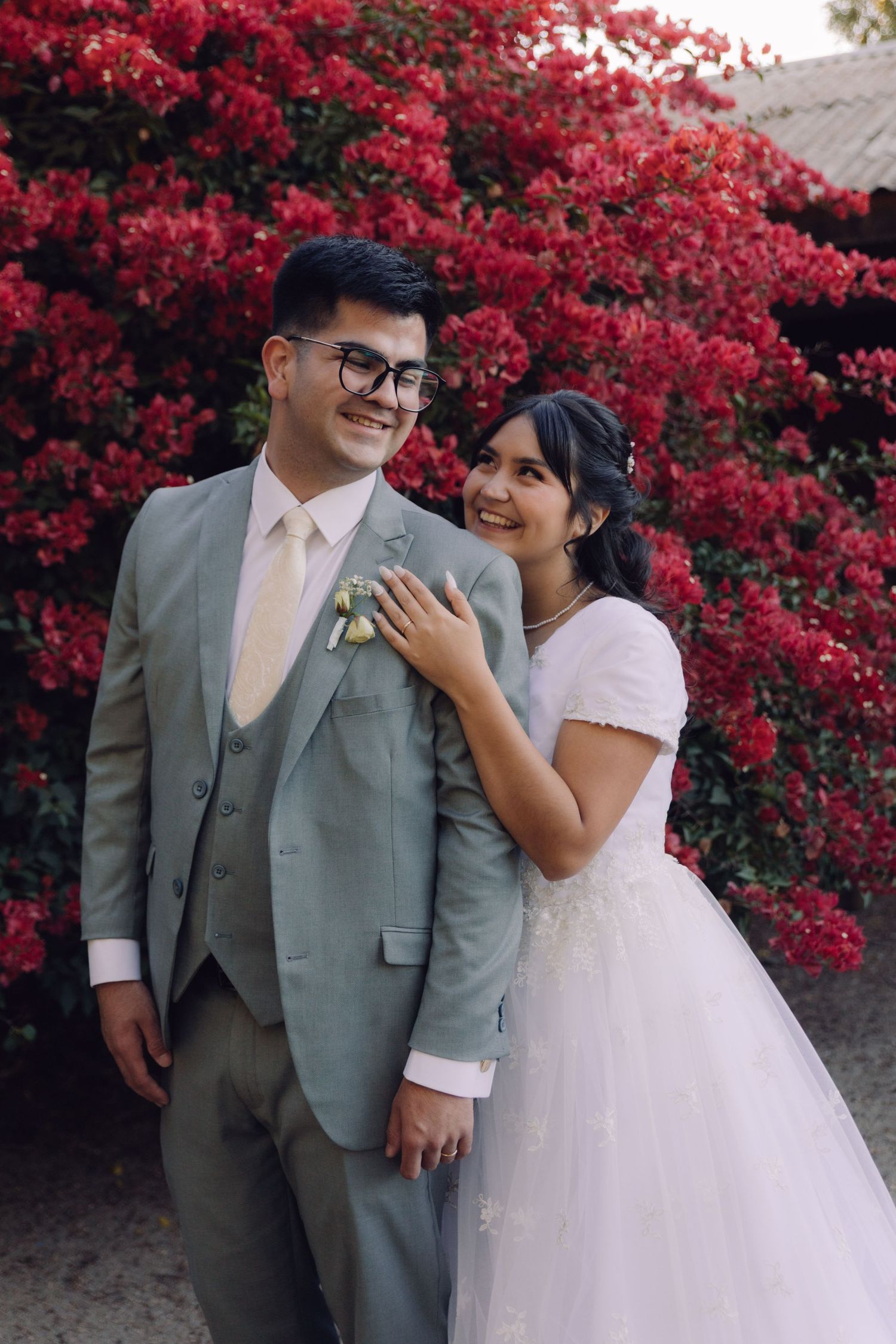 Smiling bride and groom pose together in front of bright red bougainvillea at outdoor wedding.