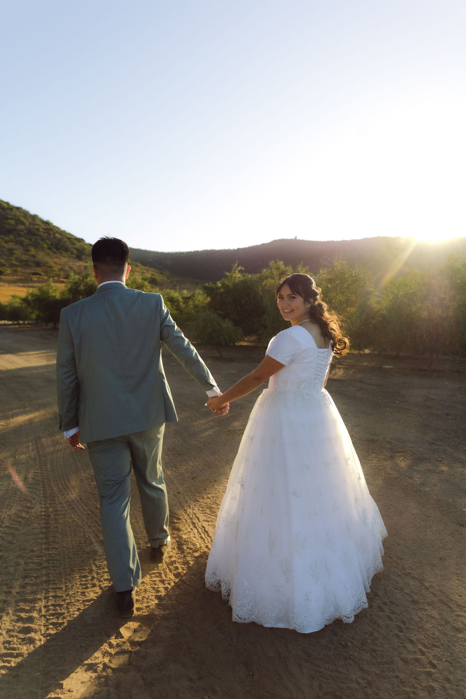 Bride and groom walk hand in hand toward golden sunset in scenic desert landscape.