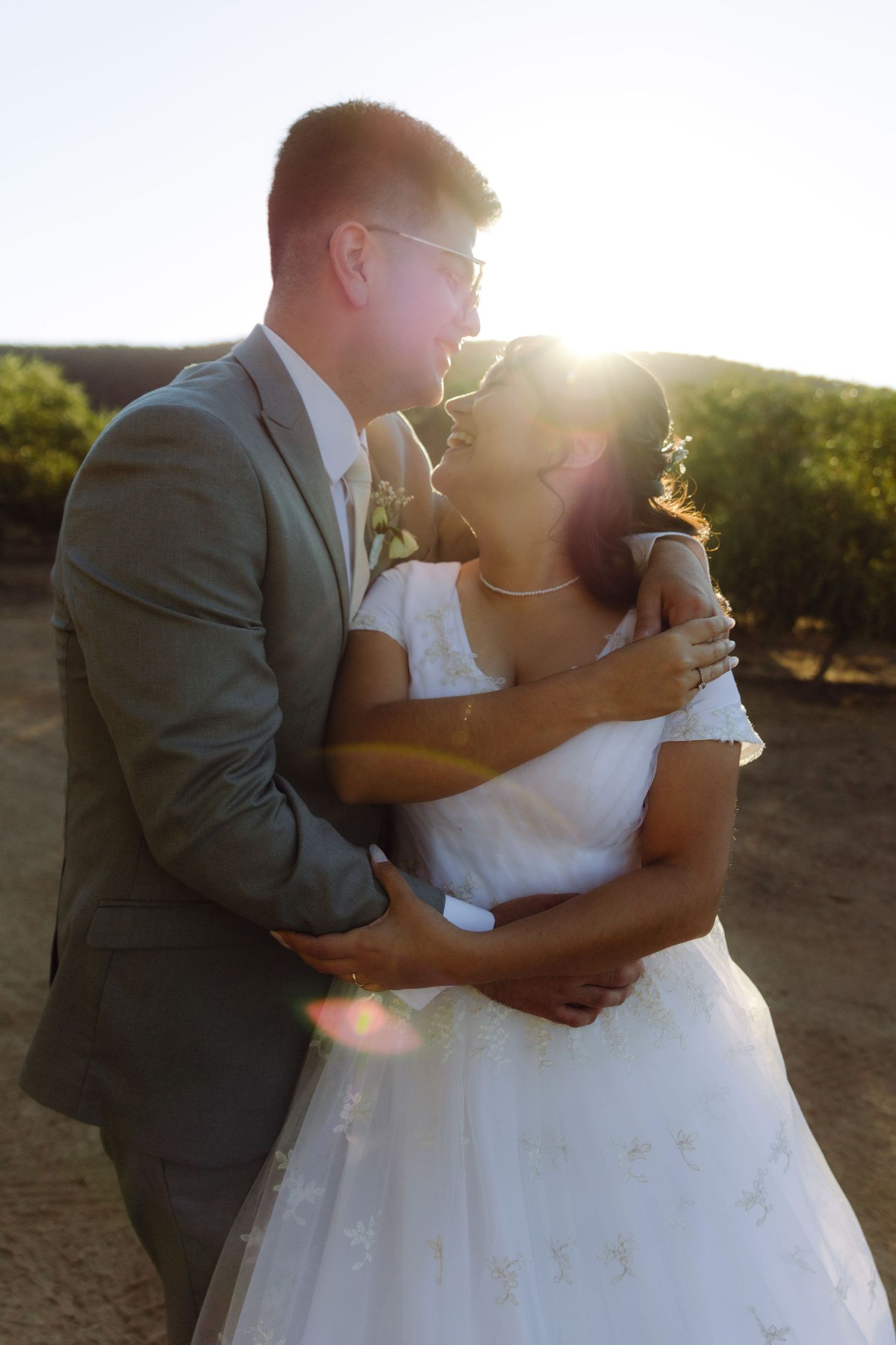 Bride and groom embrace romantically with golden sunset lens flare behind them outdoors.