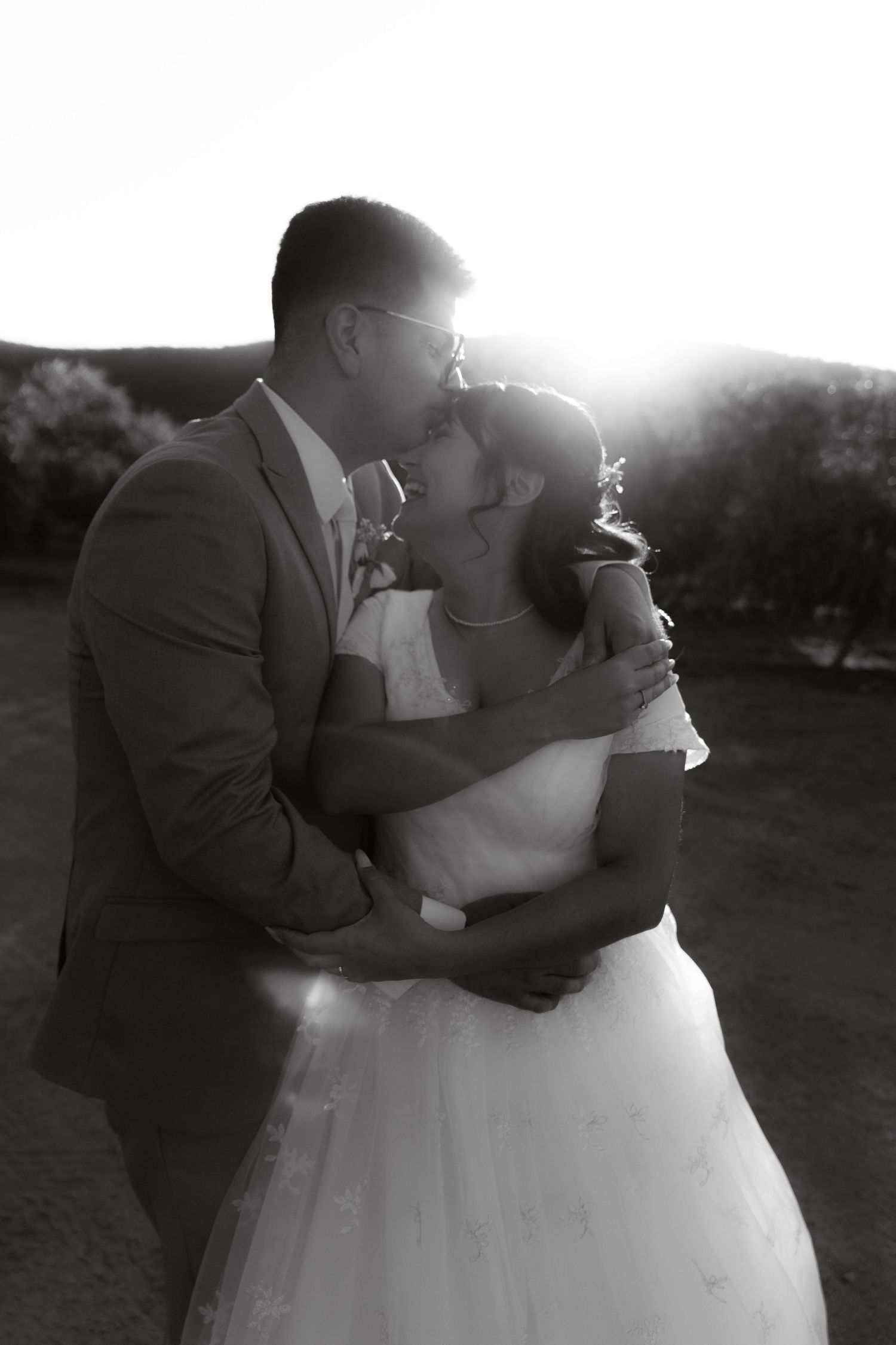 Black and white photo of bride and groom embracing with dramatic sunset backlight.